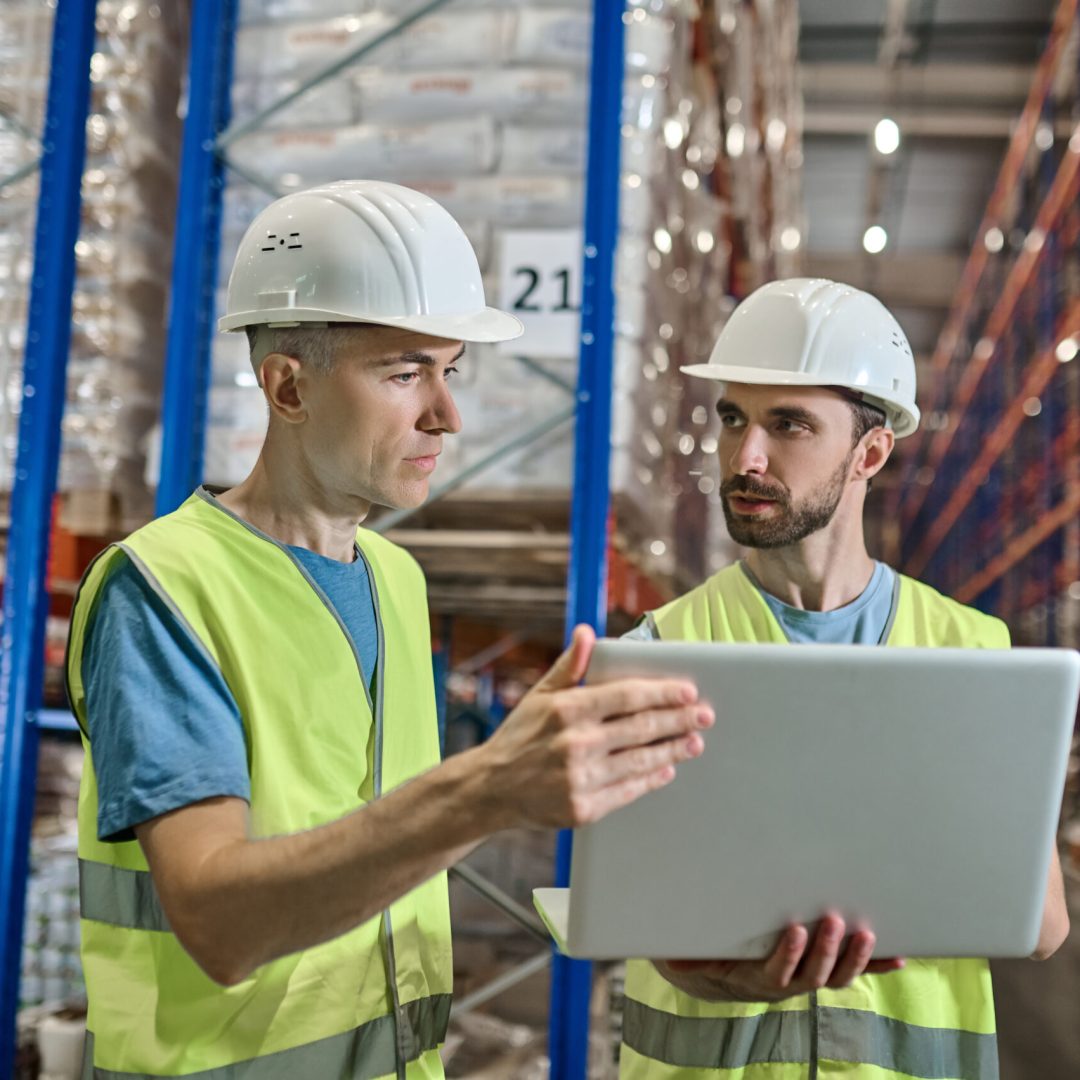 Discussion, colleagues. Two focused men in helmet and overalls standing working with open laptop discussing working moment in warehouse